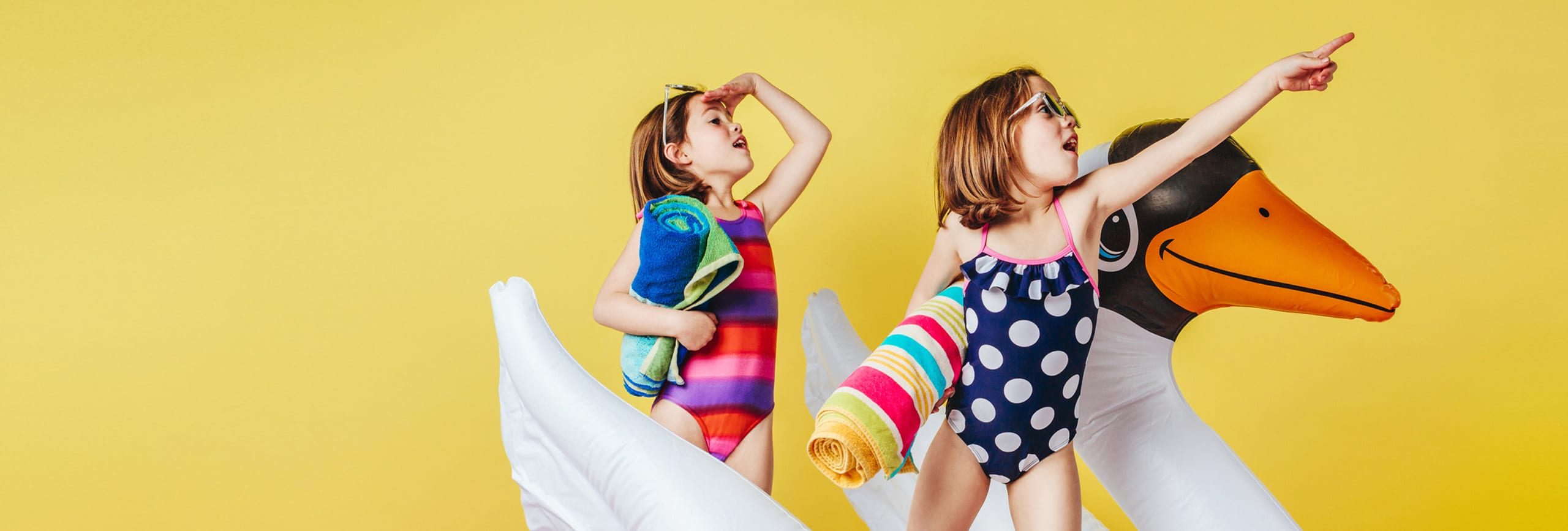 Deux petites filles en maillot de bain regardent au loin et montrent quelque chose en l'air. Elles sont des serviettes de bain et une grande bouée en forme de cygne.